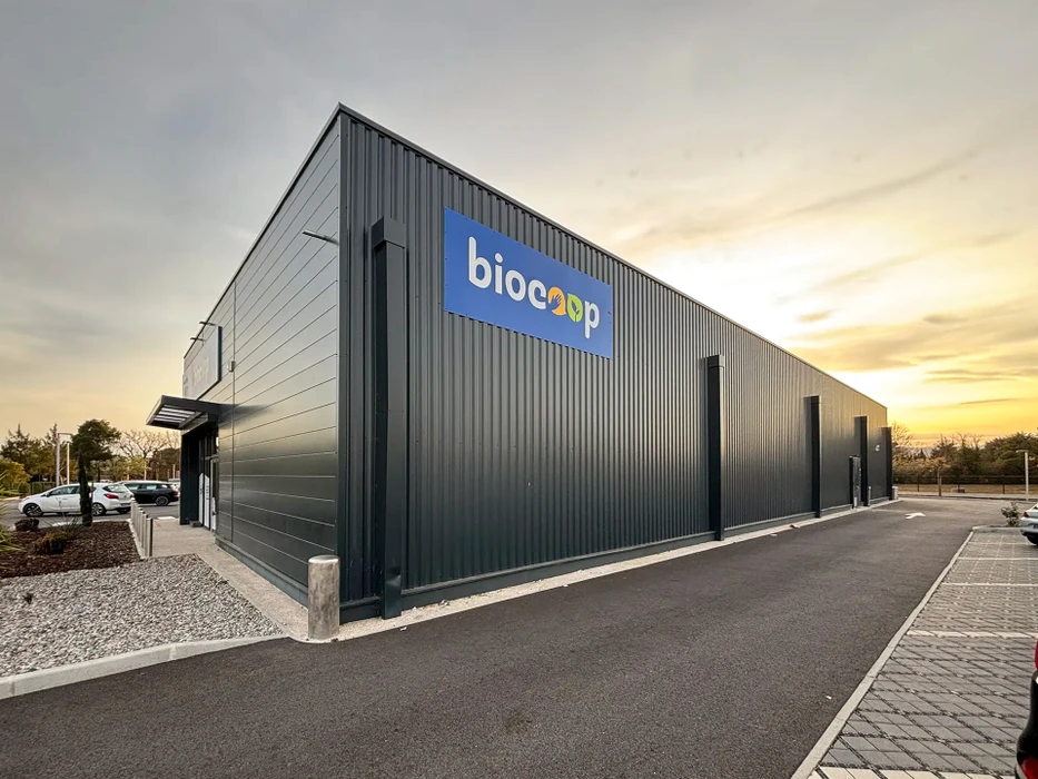 Bâtiment industriel moderne en construction sur terrain agricole, hangar métallique avec bardage en tôle ondulée gris clair, toiture à deux pans, façade percée de fenêtres rectangulaires